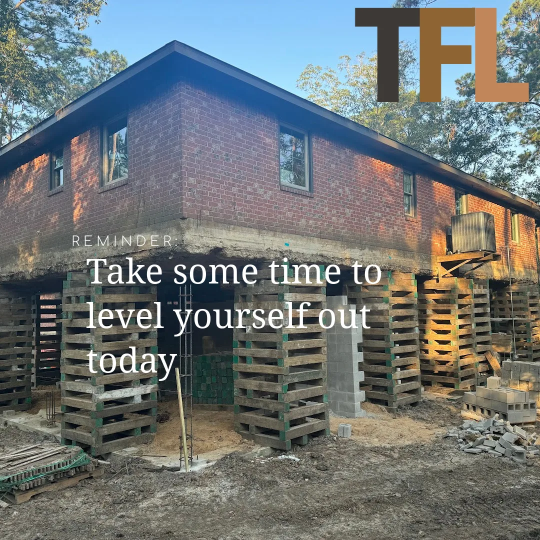 Elevated brick house supported by stacked wooden and concrete blocks during foundation repair, with trees in the background and construction materials on site.